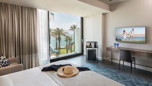 Quarto de hotel moderno com uma varanda grande, uma cama, uma escrivaninha e uma televisão exibindo uma cena de praia.