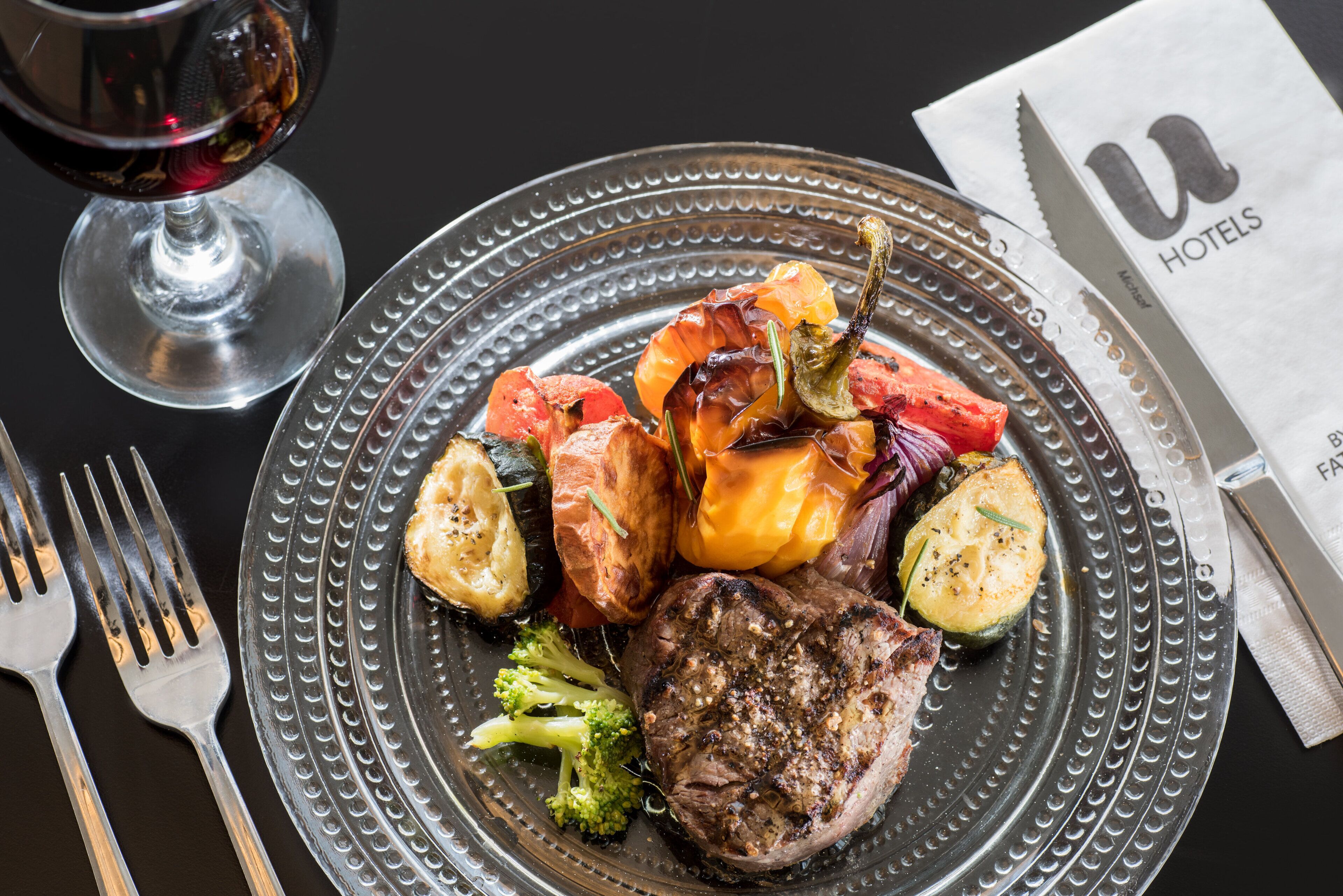 A plate of food with steak, vegetables, and a glass of red wine on a table.
