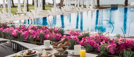 Una mesa de comedor con alimentos para el desayuno, una piscina y flores rosadas al fondo.