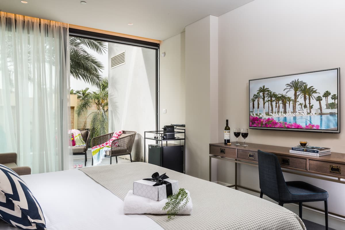 Ein modernes Hotelzimmer mit Bett, Schreibtisch, Sessel, einem Gemälde und Blick auf Palmen und einen Pool.