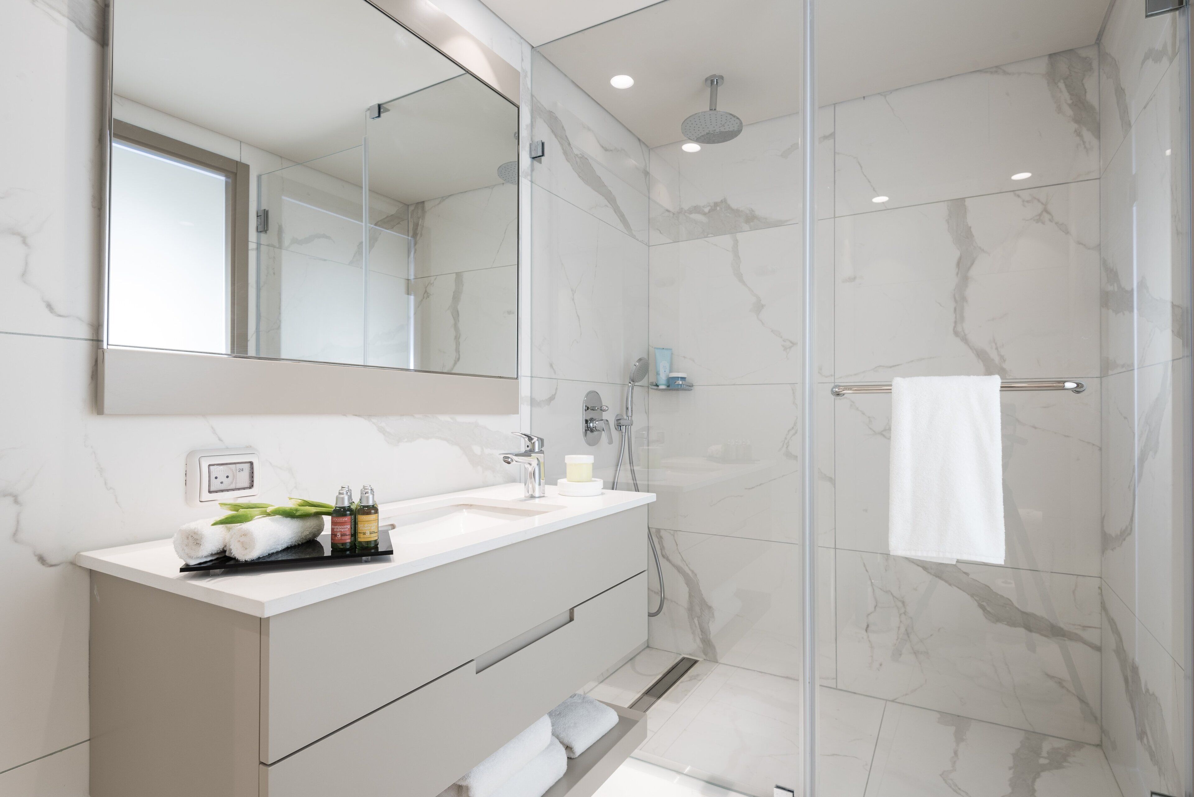 A modern bathroom with a glass-enclosed shower, a white vanity with a mirror, and marble walls.