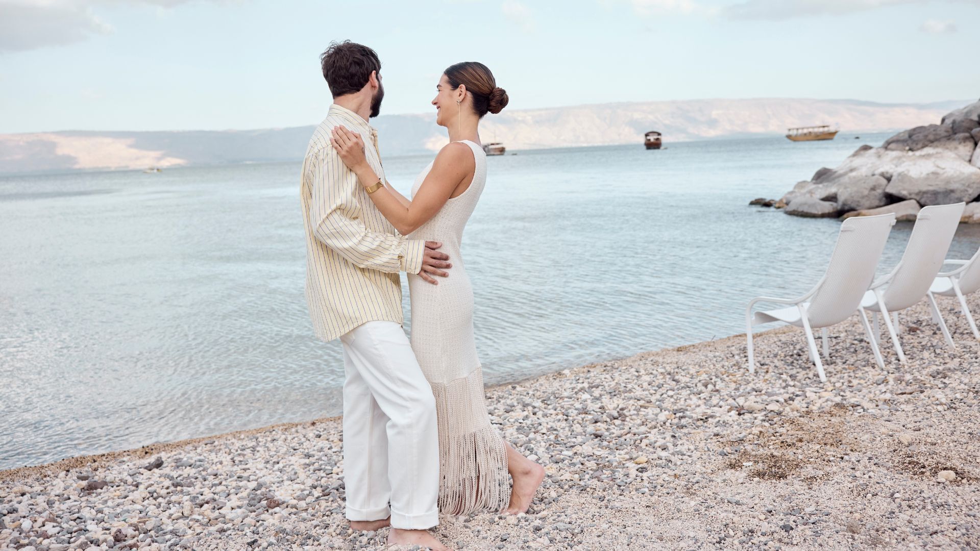 A couple standing on a pebbled beach with a calm sea and boats in the distance.