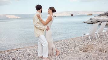 A couple standing on a pebbled beach with a calm sea and boats in the distance.