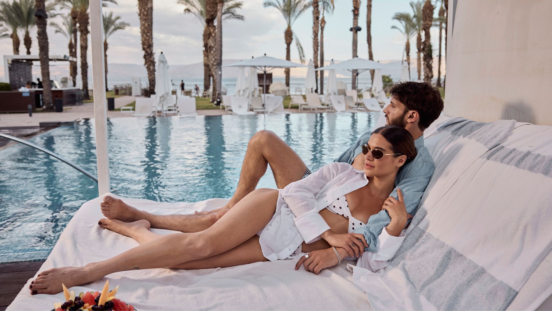 Una pareja descansando en una silla de piscina con vista a la alberca y palmeras.