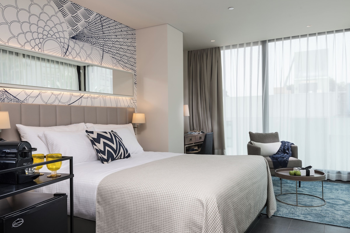 A modern hotel room with a large bed, a desk with a coffee maker, a chair, and a view of the city through large windows.