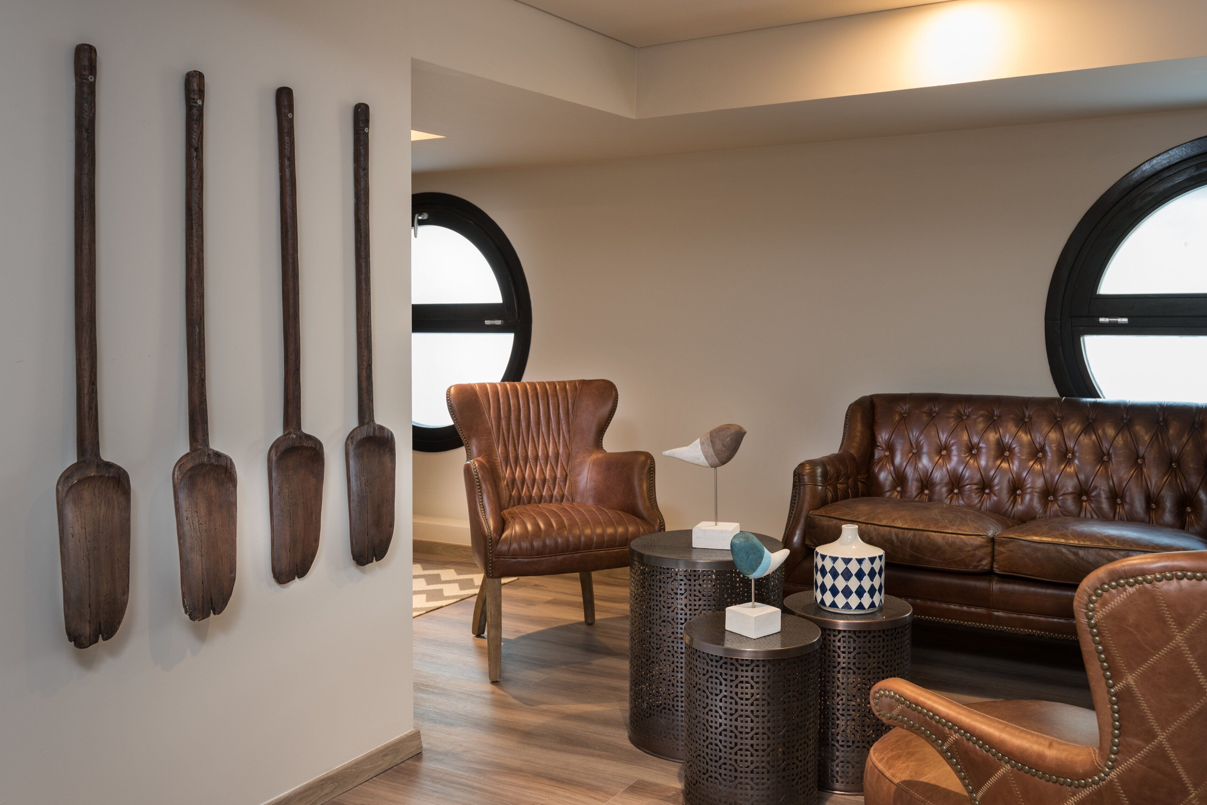 Una sala moderna con un sillón de cuero, un sofá capitonado y una mesa de centro. Hay cuatro palas de madera montadas en la pared y dos mesas redondas con objetos decorativos.