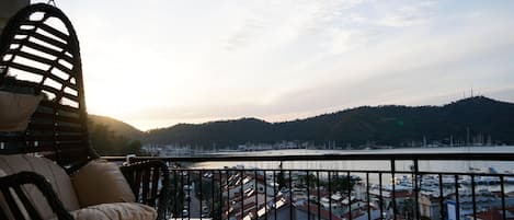A balcony with a wicker chair and a view of a marina with boats and a hilly landscape in the background.