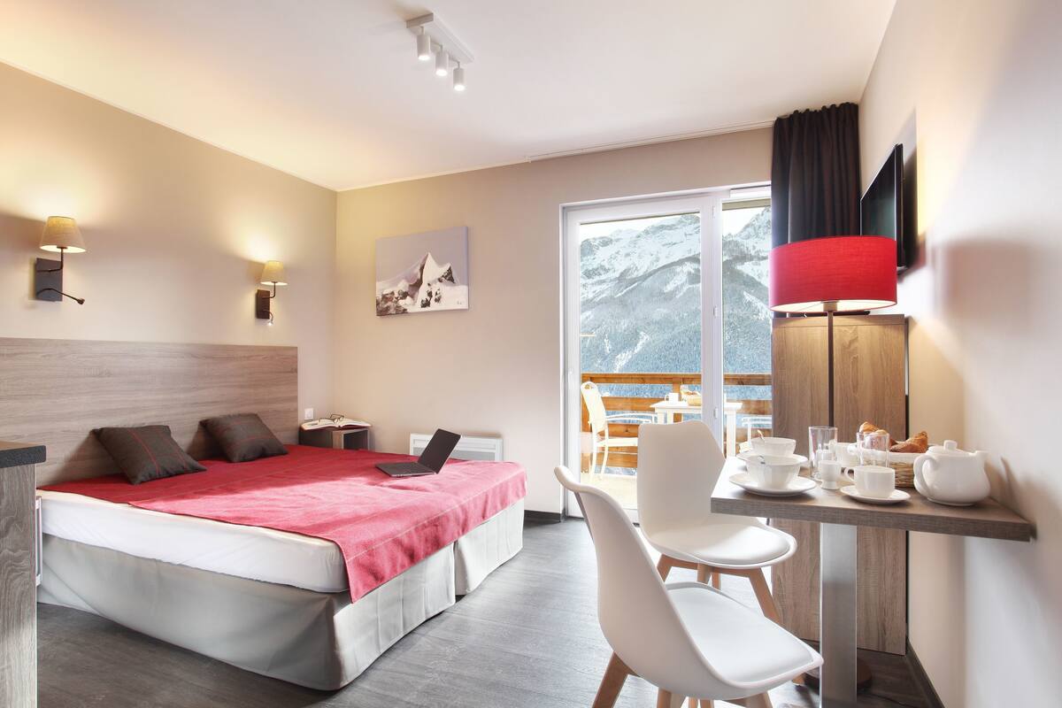 A hotel room with a bed, a desk with a laptop, a chair, and a table with breakfast items.