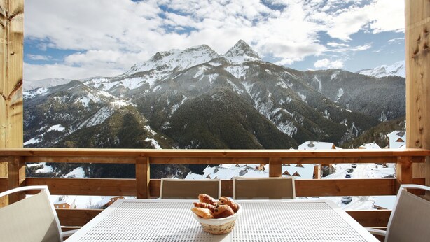 Une terrasse en bois avec une table et des chaises, un panier de croissants et une vue sur la montagne.