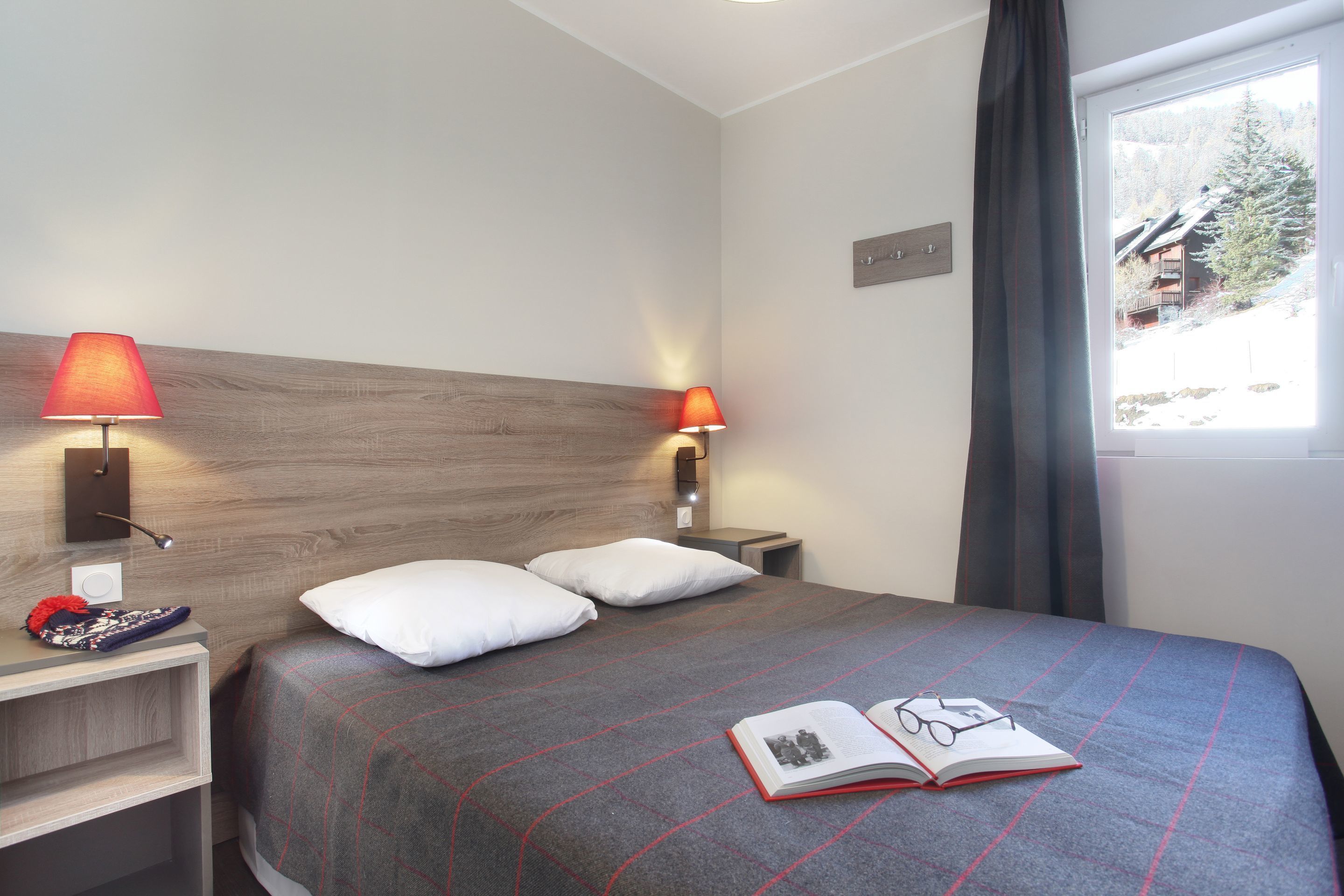 Habitación de hotel con cama, dos lámparas de noche, una mesita de noche y ventana con vistas a un edificio y árboles.