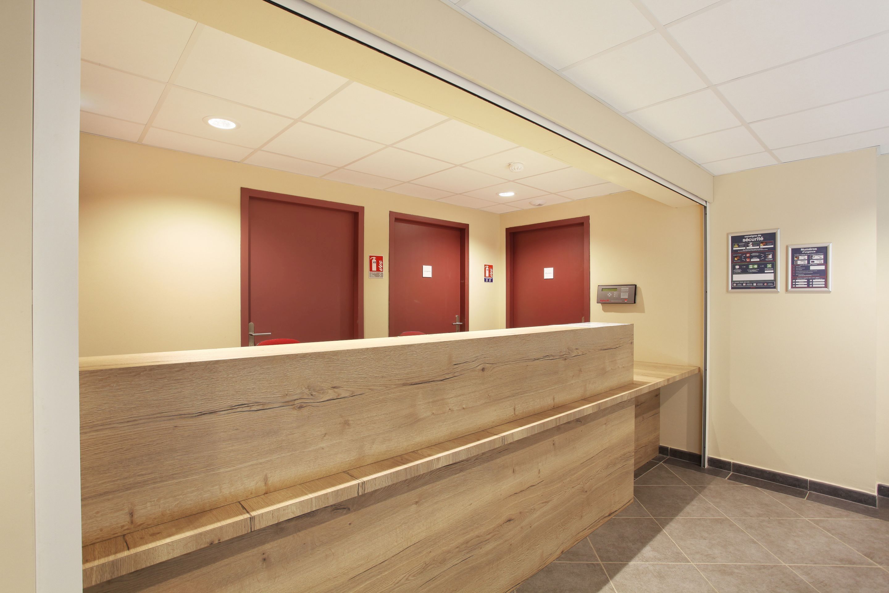 Un espace d’accueil avec un comptoir de réception en bois, trois portes rouges et un sol carrelé.