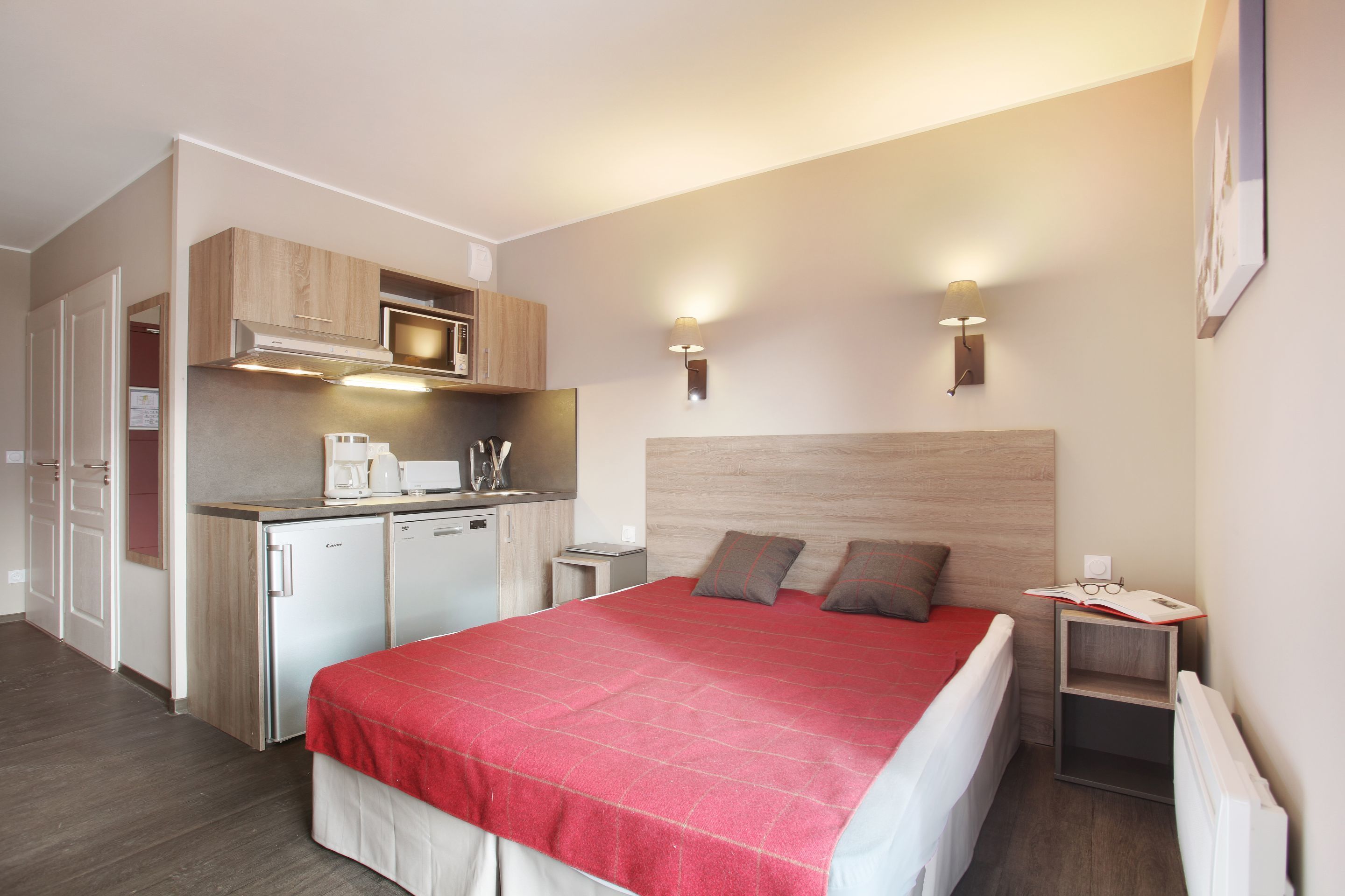 A compact hotel room with a bed, a kitchenette, and a small bedside table.