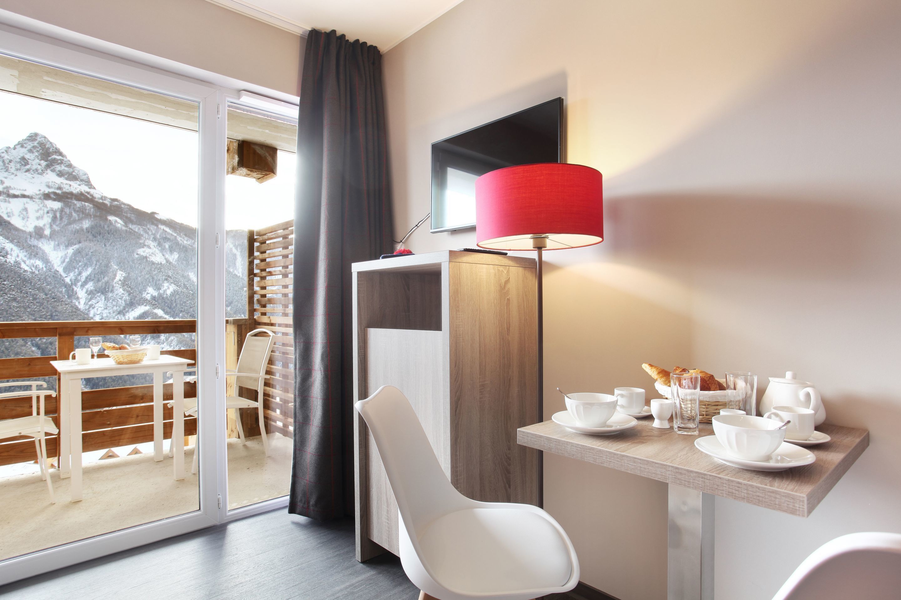 Una camera d'albergo moderna con balcone, un tavolo da pranzo apparecchiato con stoviglie bianche, una lampada rossa e una TV a schermo piatto appesa al muro.