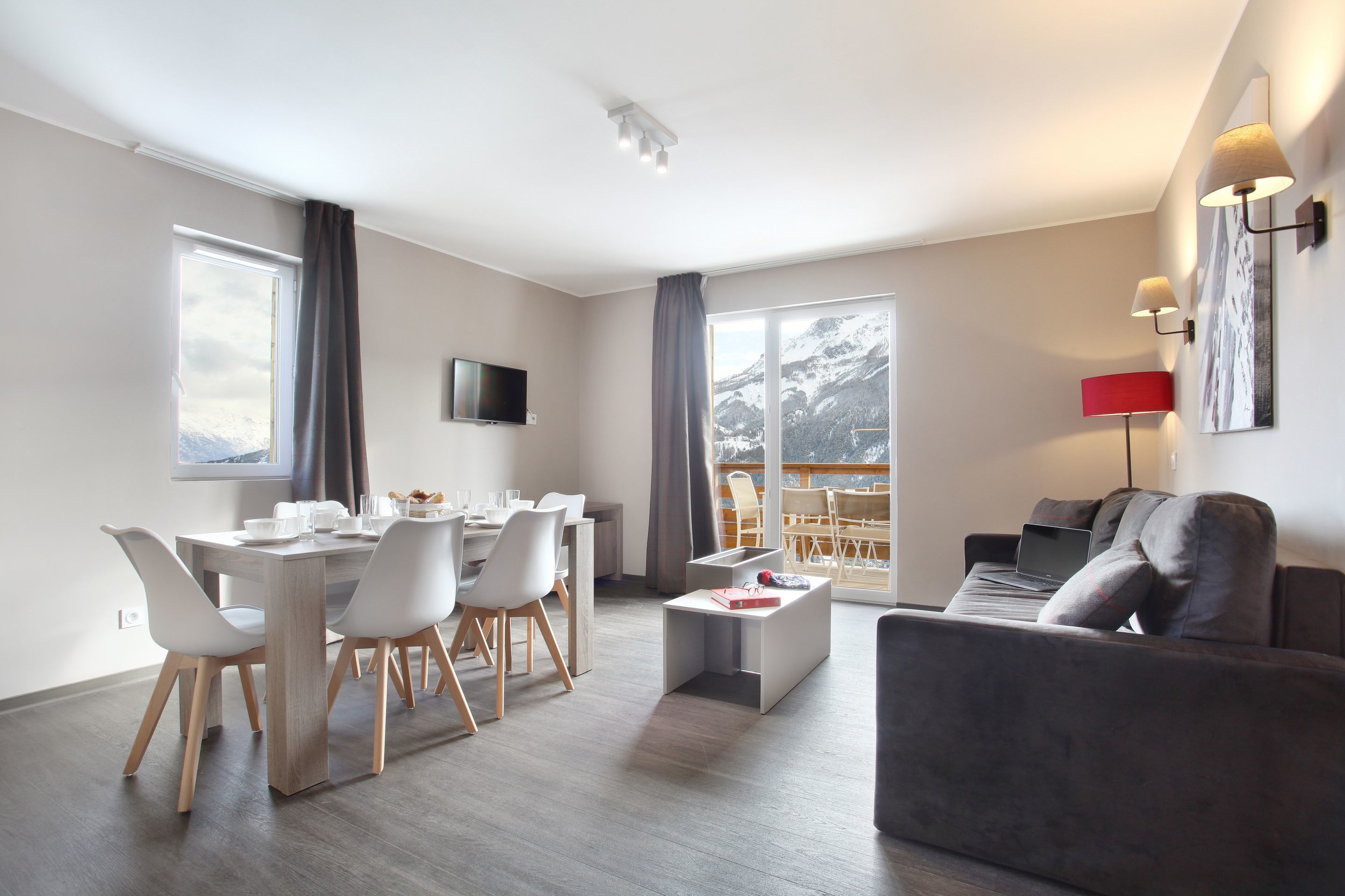 A modern living room with a grey sofa, a dining area with white chairs, a flat-screen TV mounted on the wall, and a large window with a view of snow-capped mountains.
