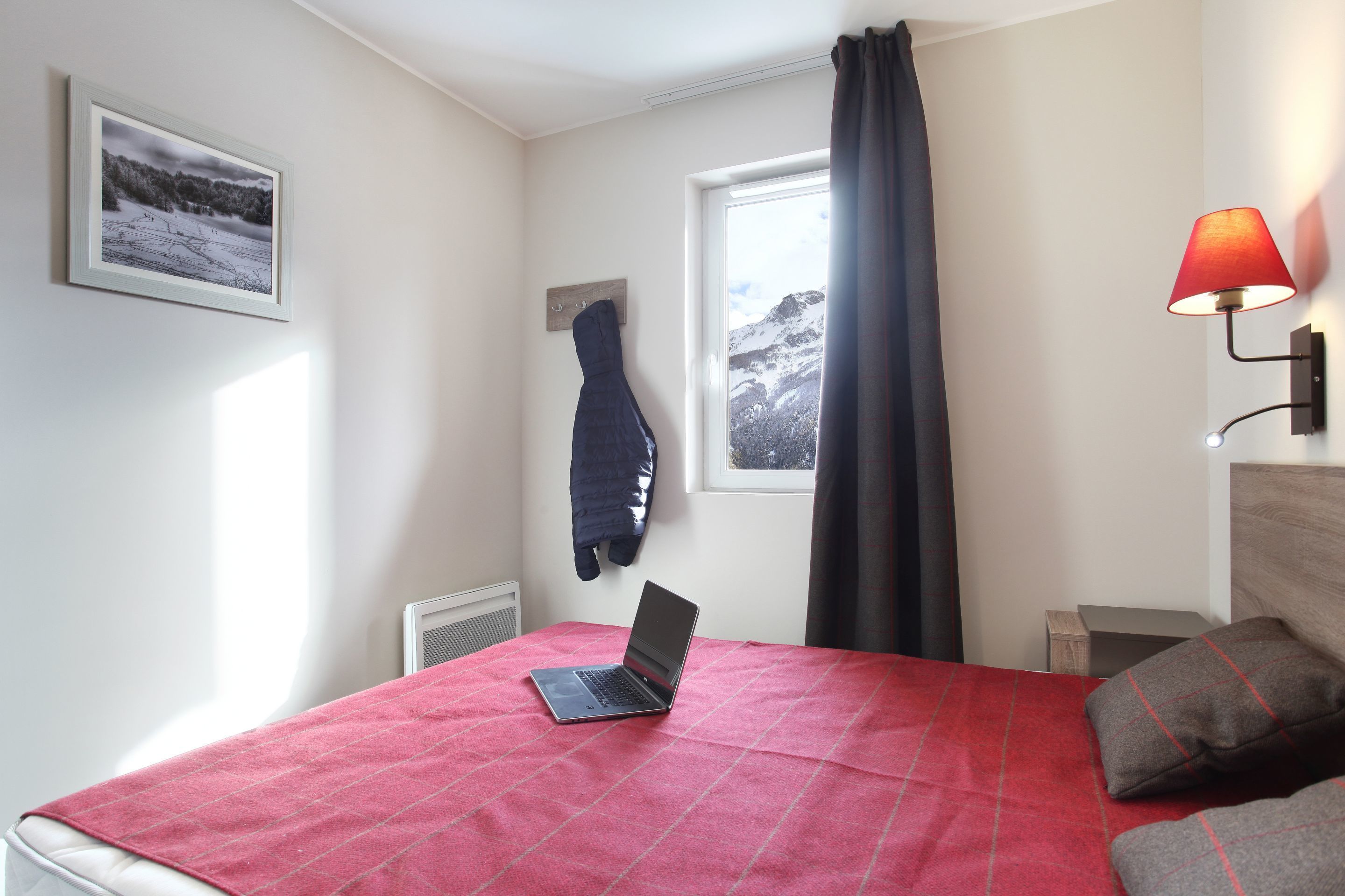 Une chambre avec un ordinateur portable posé sur le lit, une fenêtre donnant sur les montagnes, une couverture rouge et une lampe fixée au mur.