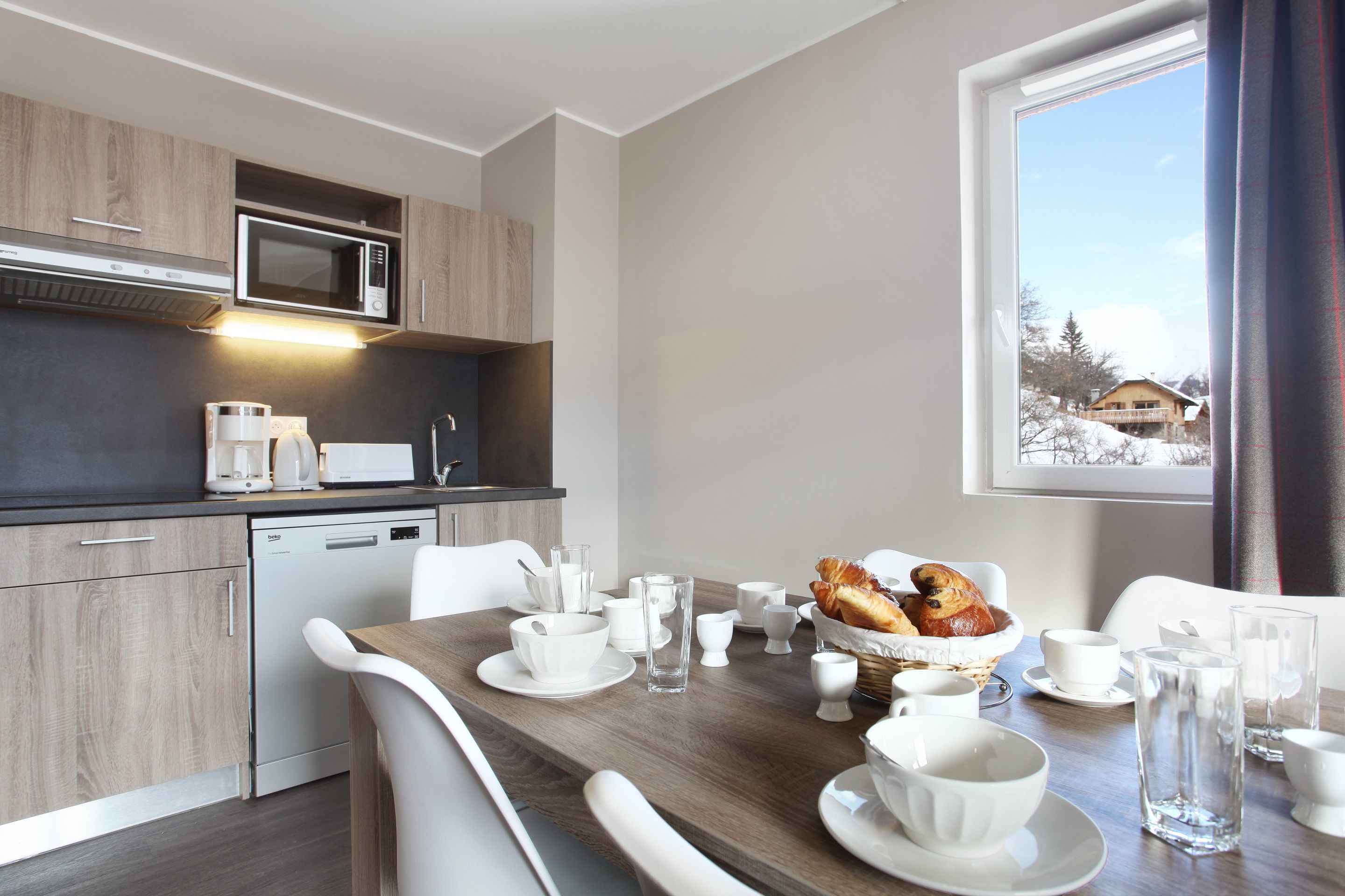 A modern kitchen with a microwave, coffee maker, and dishwasher. There is a dining table with white chairs and a basket of bread.
