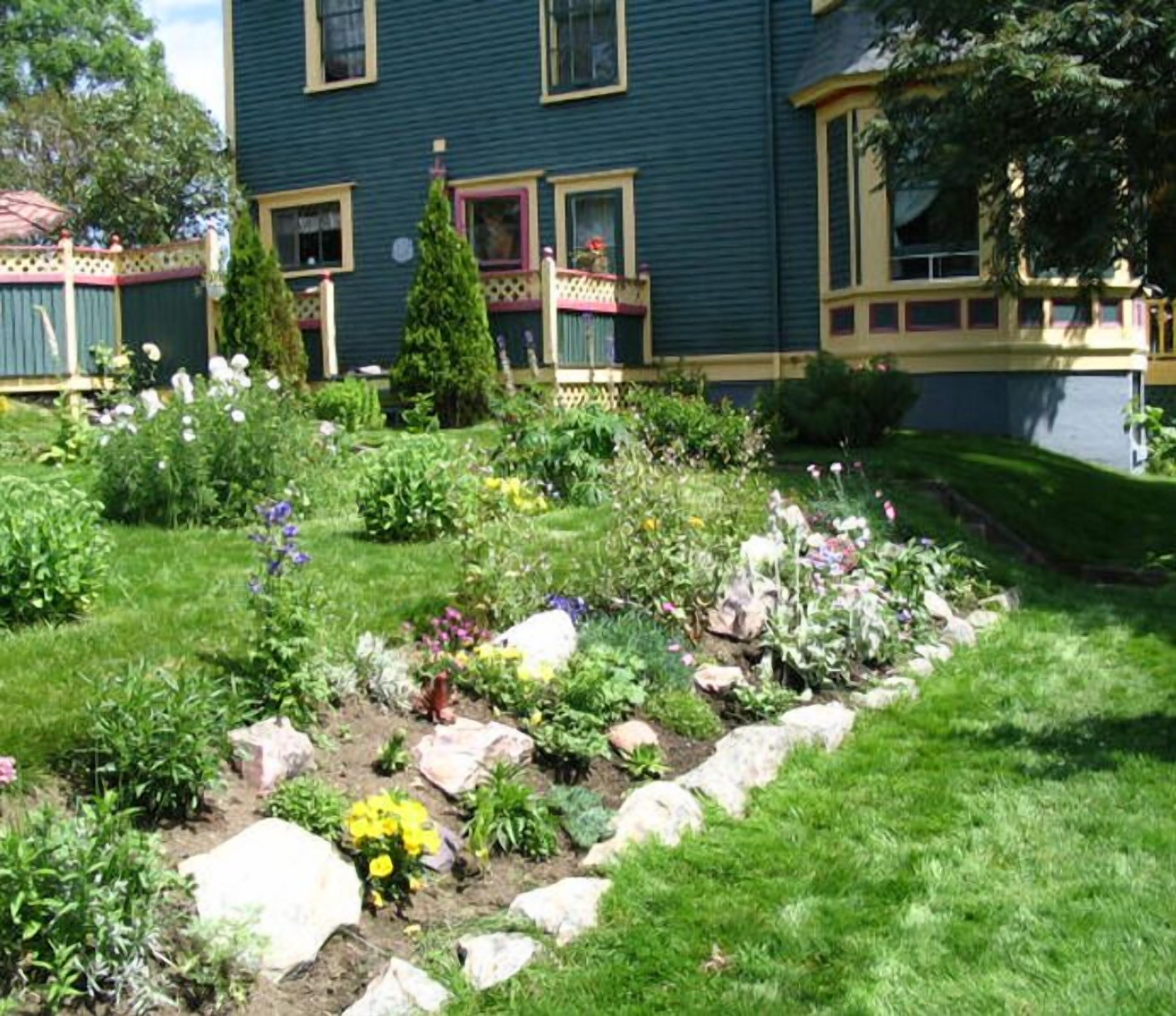 En have med forskellige planter og blomster foran et hus med blå facade og gul detaljering.
