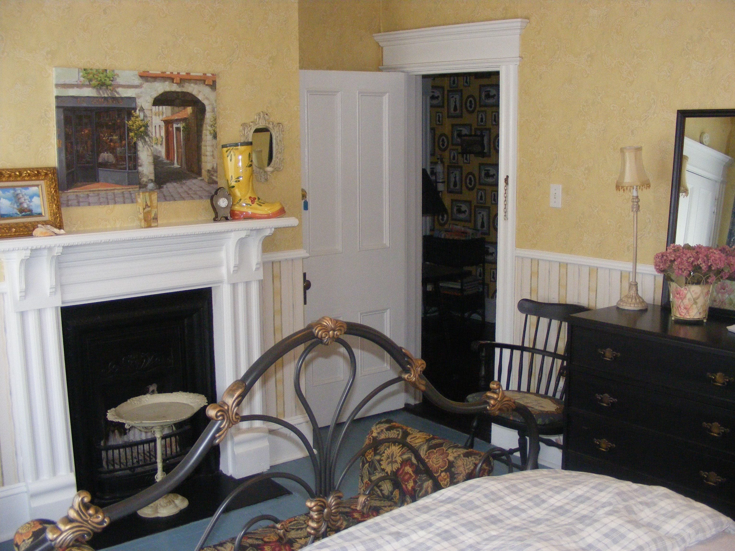 A bedroom with a fireplace, a bed, a dresser, a chair, and a door leading to a hallway.