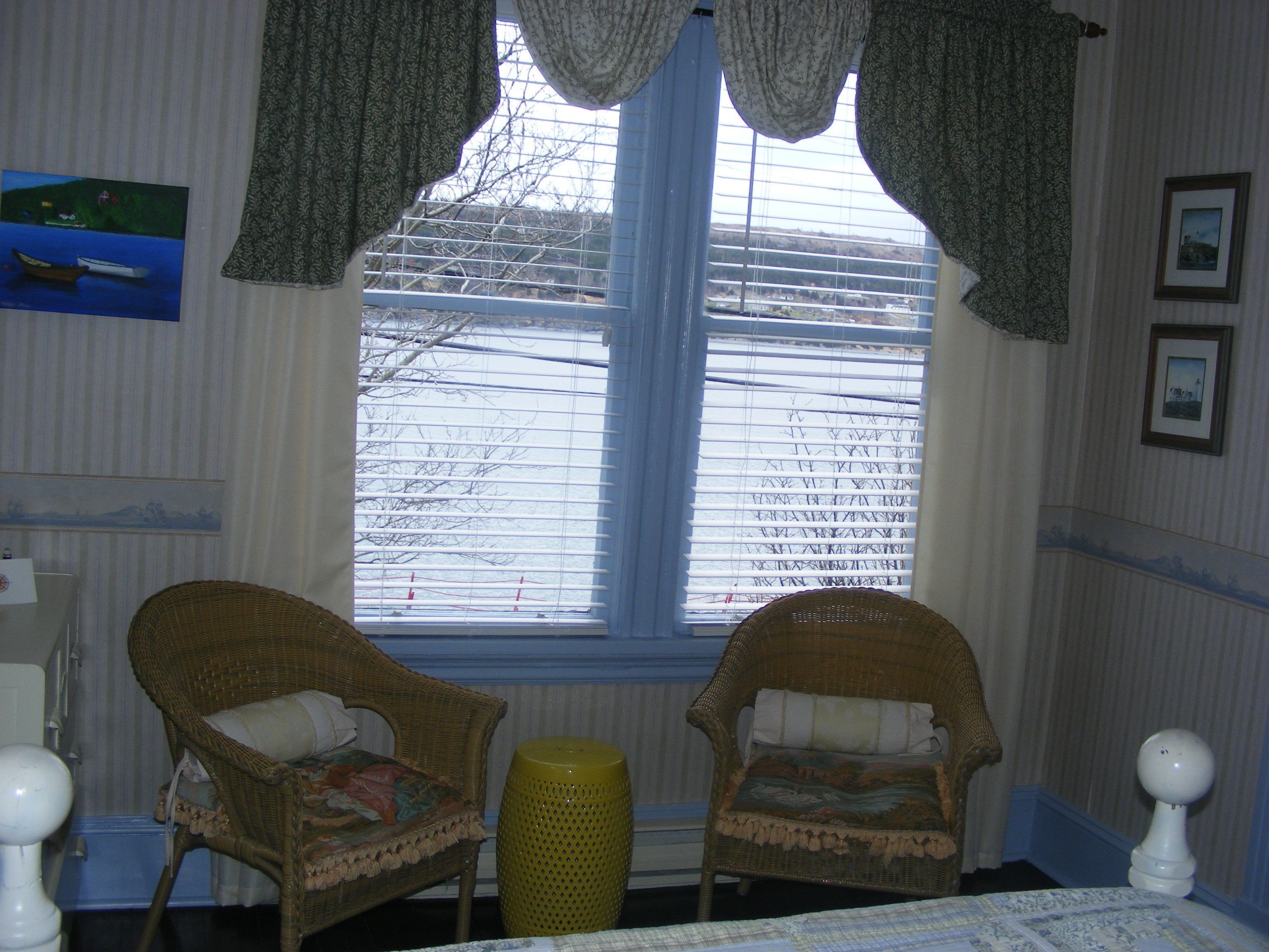 A room with two wicker chairs, a bed with a patterned bedspread, a window with blinds, and a small yellow stool.