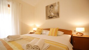 Ein ordentlich bezogenes Bett mit weißen und gestreiften Kissen, einer gesteppten Tagesdecke und aufgerollten Handtüchern. Darüber an der Wand hängt ein Gemälde, und auf den Holz Nachttischen stehen je eine Nachttischlampe.