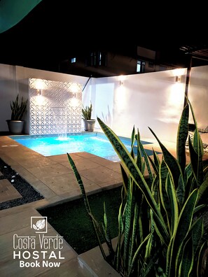 Un área de piscina con un diseño de pared geométrico, plantas en macetas y un letrero de Costa Verde HOSTAL.