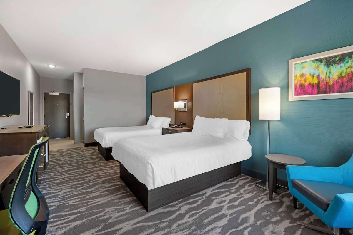 A hotel room with two beds, a desk, a chair, a TV, a lamp, and a colorful painting on the wall.