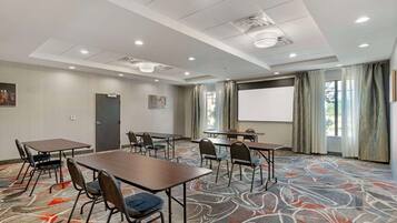 A conference room with tables, chairs, a projector screen, and a large window with curtains.