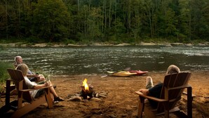Two people sitting on wooden chairs by a campfire, with a river and forest in the background. - Paddler's Lane Retreat (Confluence)