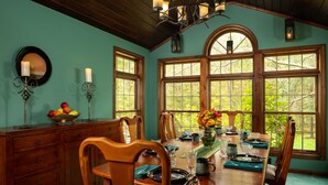 A dining room with a wooden table set for four, surrounded by chairs, with a large window providing a view of greenery. - Paddler's Lane Retreat (Confluence)