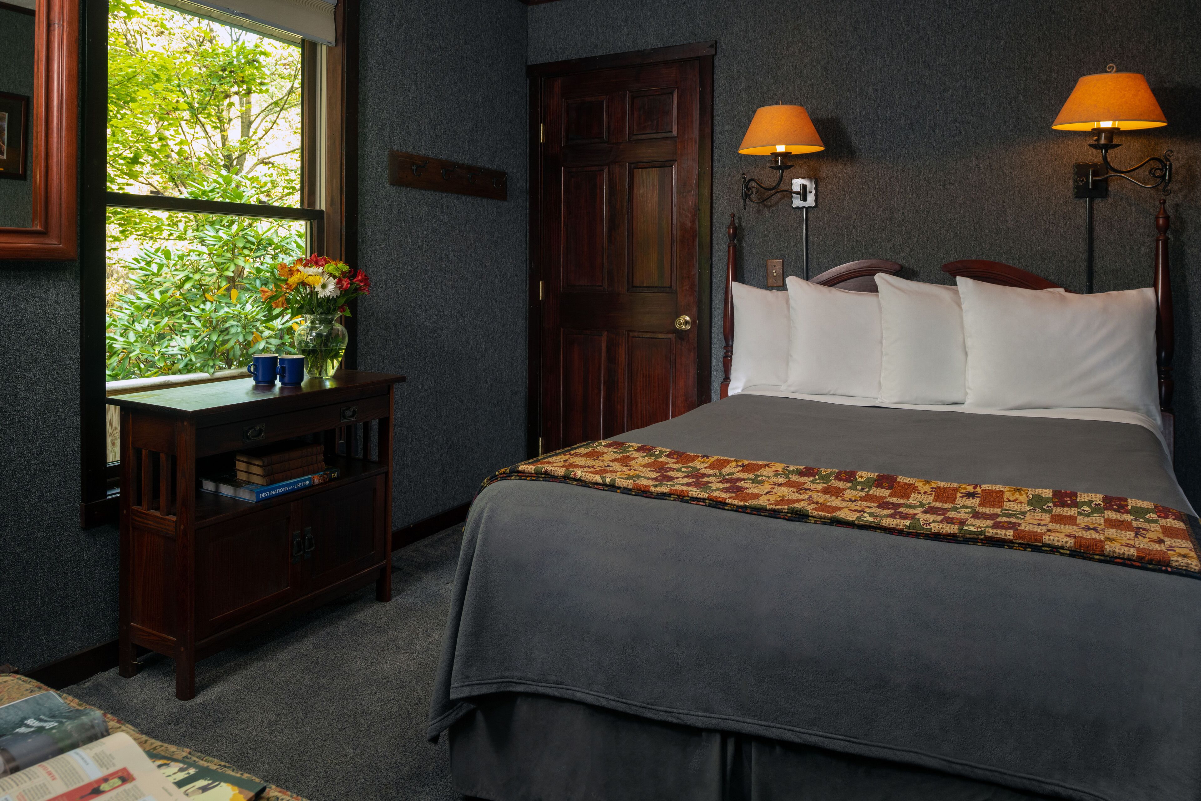 Un dormitorio con una cama grande, dos lámparas de noche, una ventana con vistas a la vegetación, una mesita de noche de madera con libros y un jarrón de flores, y suelo alfombrado.