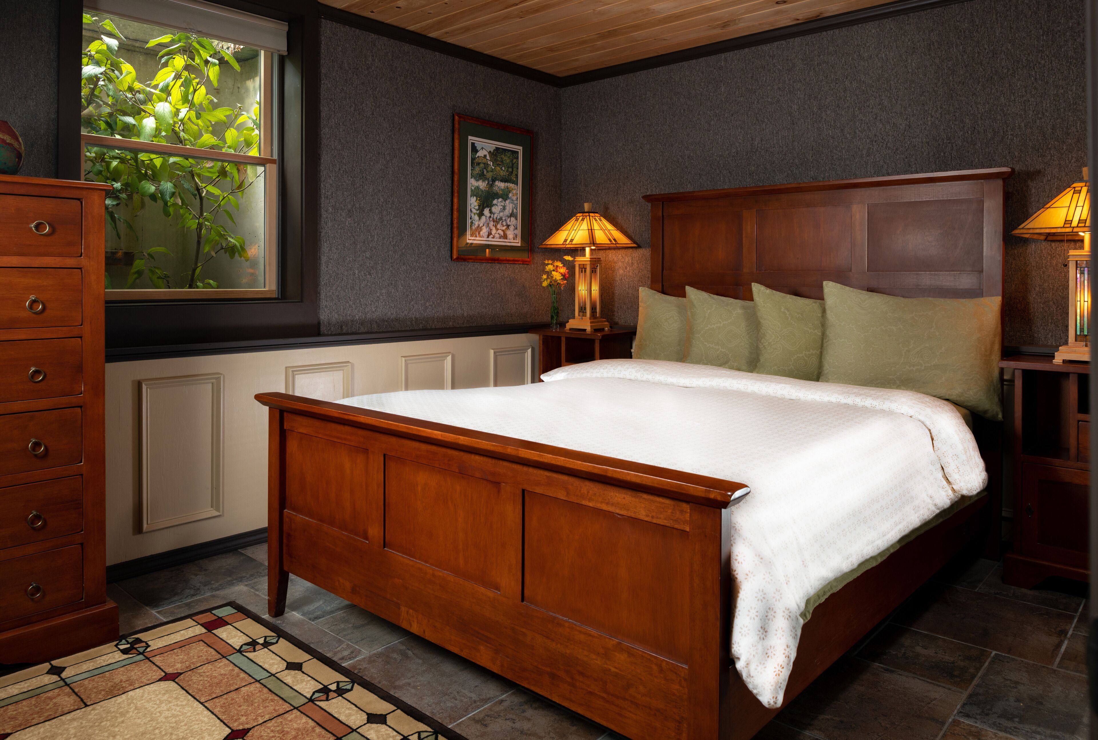 A bedroom with a wooden bed, two bedside lamps, a dresser, a window with a view of greenery, and a framed picture on the wall.