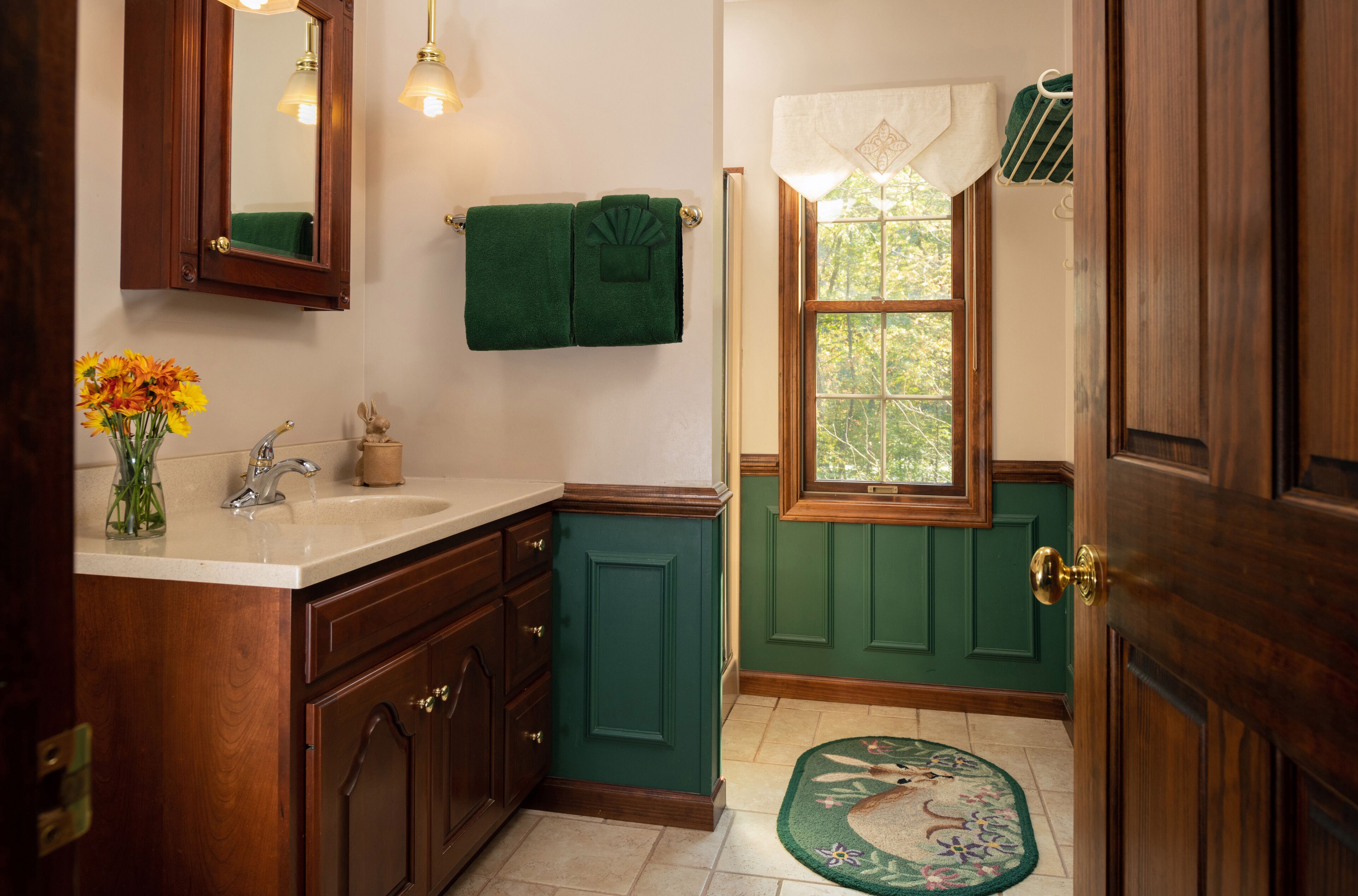 A bathroom with a wooden vanity, green walls, a window with a view, and a green towel on the wall.