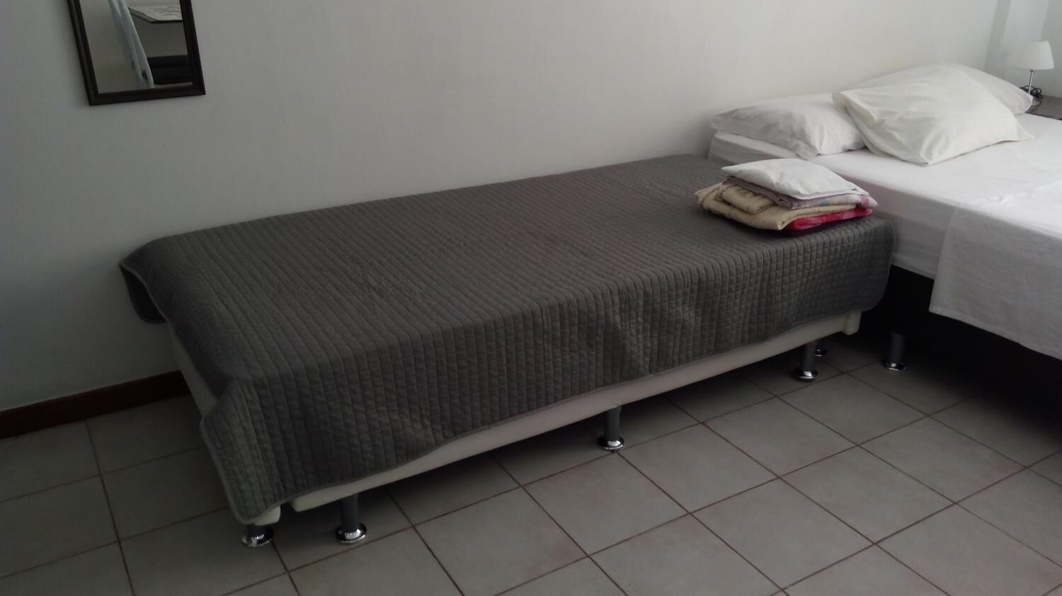 A single bed with a grey quilt, a small bedside table with a lamp, and a mirror on the wall.