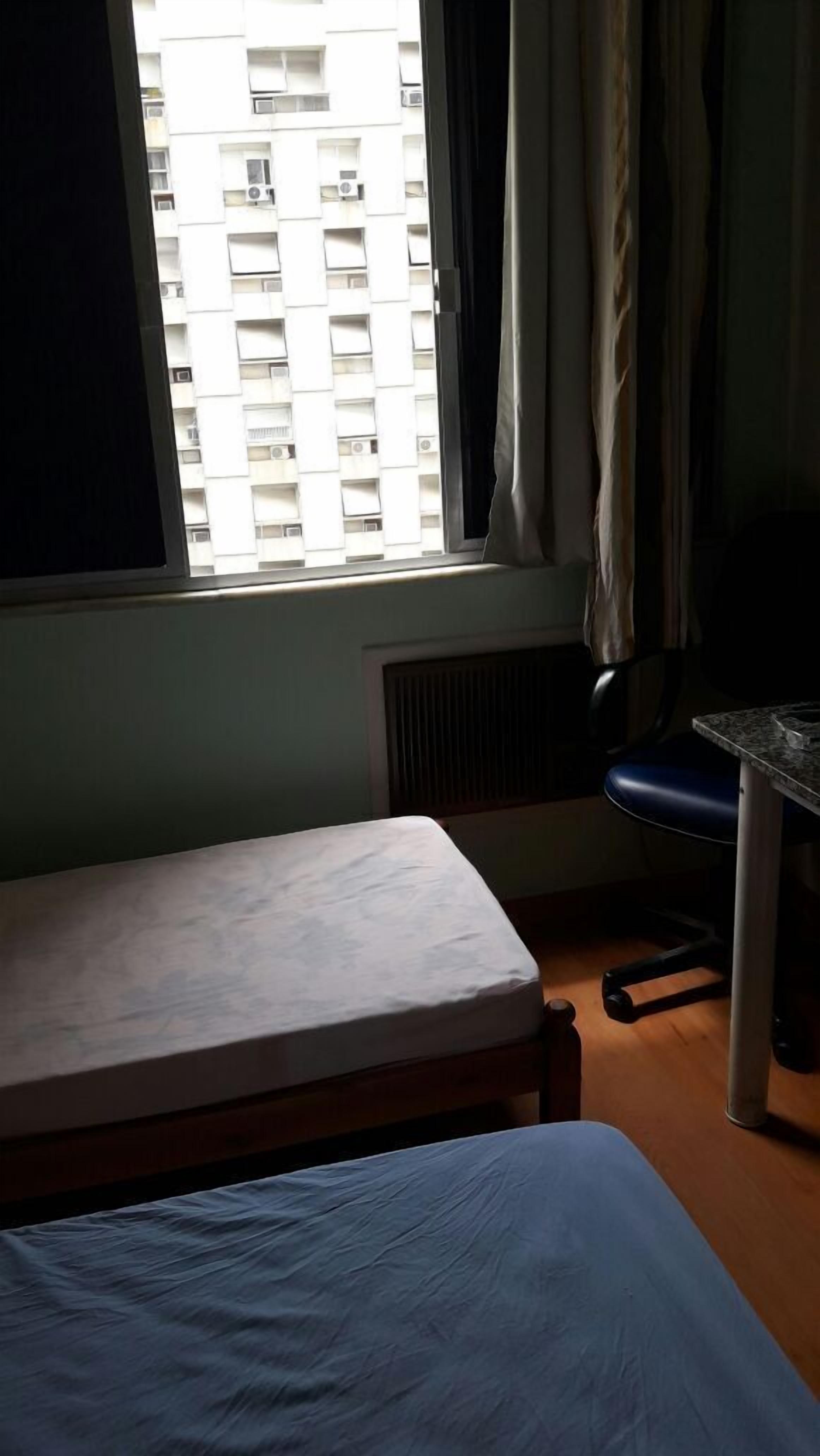 Quarto com cama, escrivaninha e cadeira. Há uma janela com cortinas, e um edifício visível lá fora.