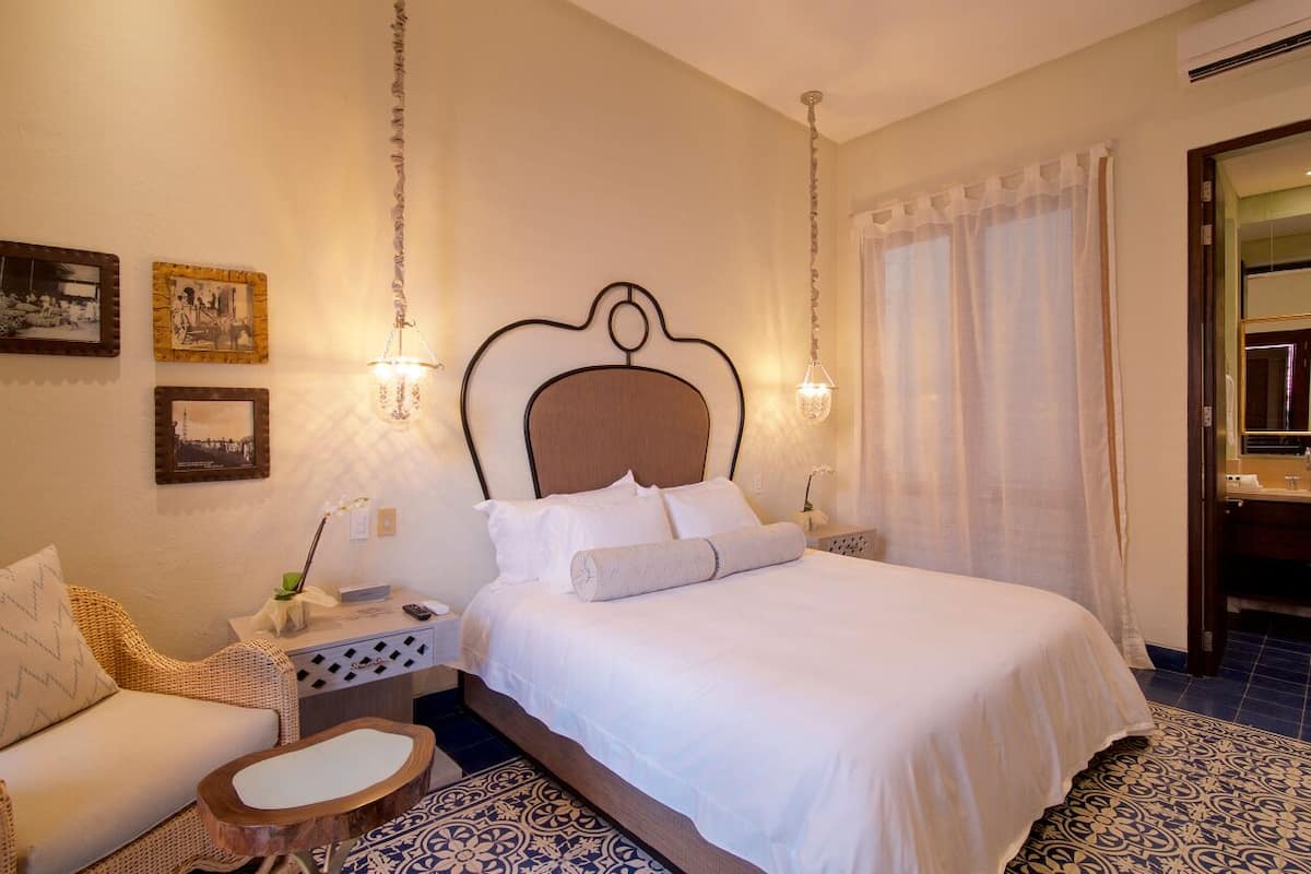 Un dormitorio con cama, mesita de noche, una silla, una mesita auxiliar de madera y una alfombra estampada.