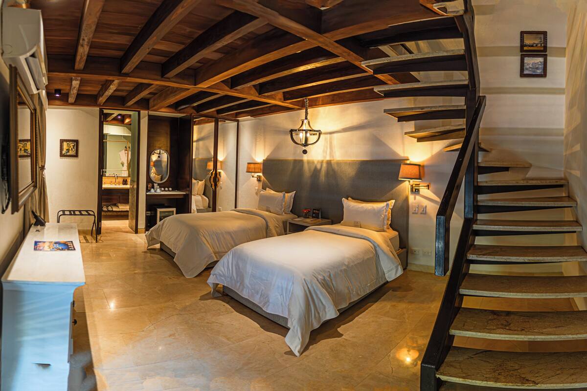 Habitación de hotel con dos camas, una escalera de madera y un baño con espejo y lavamanos.