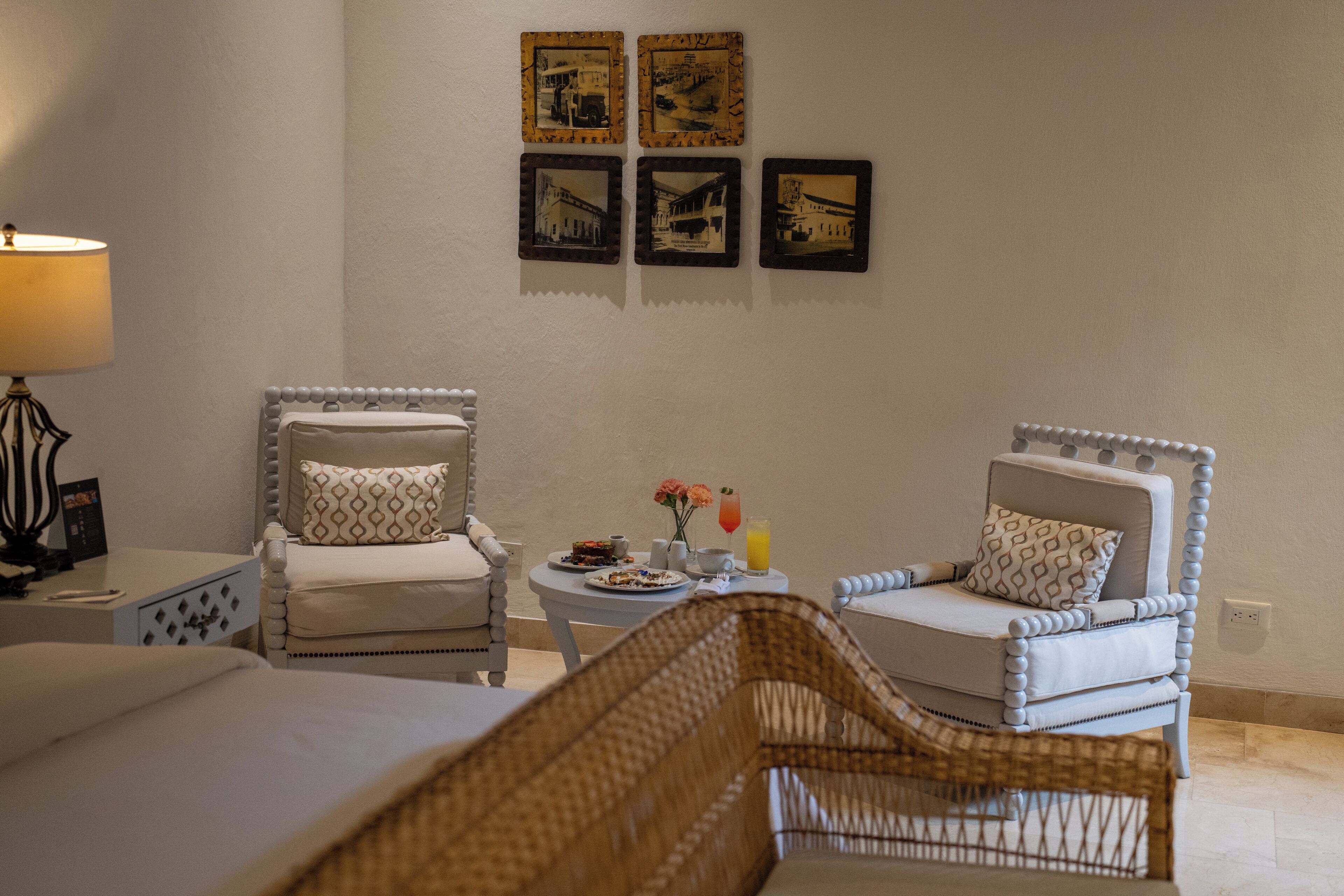 Una habitación de hotel con dos sillones tapizados, una mesita redonda con una bebida y un plato de comida, y una cama con edredón blanco.