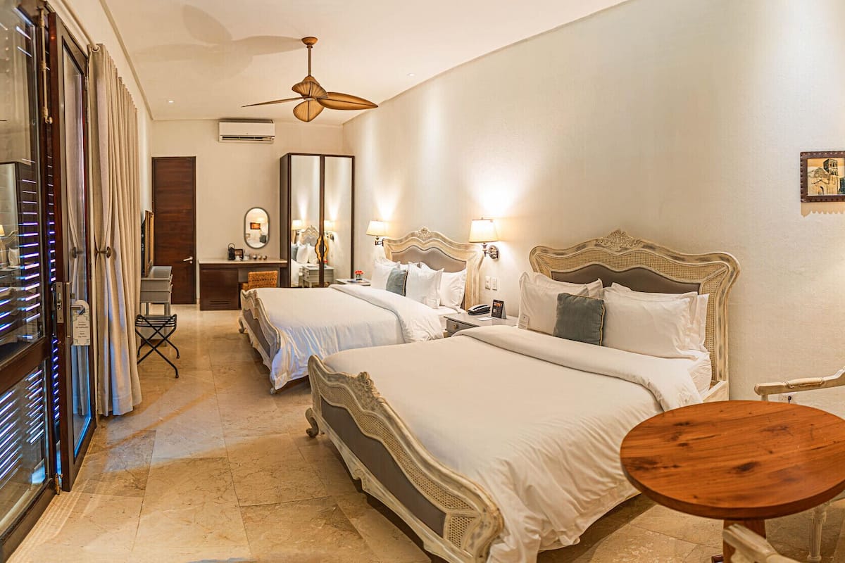 Habitación de hotel con dos camas, un tocador, un espejo, un ventilador de techo y una pequeña mesa redonda.