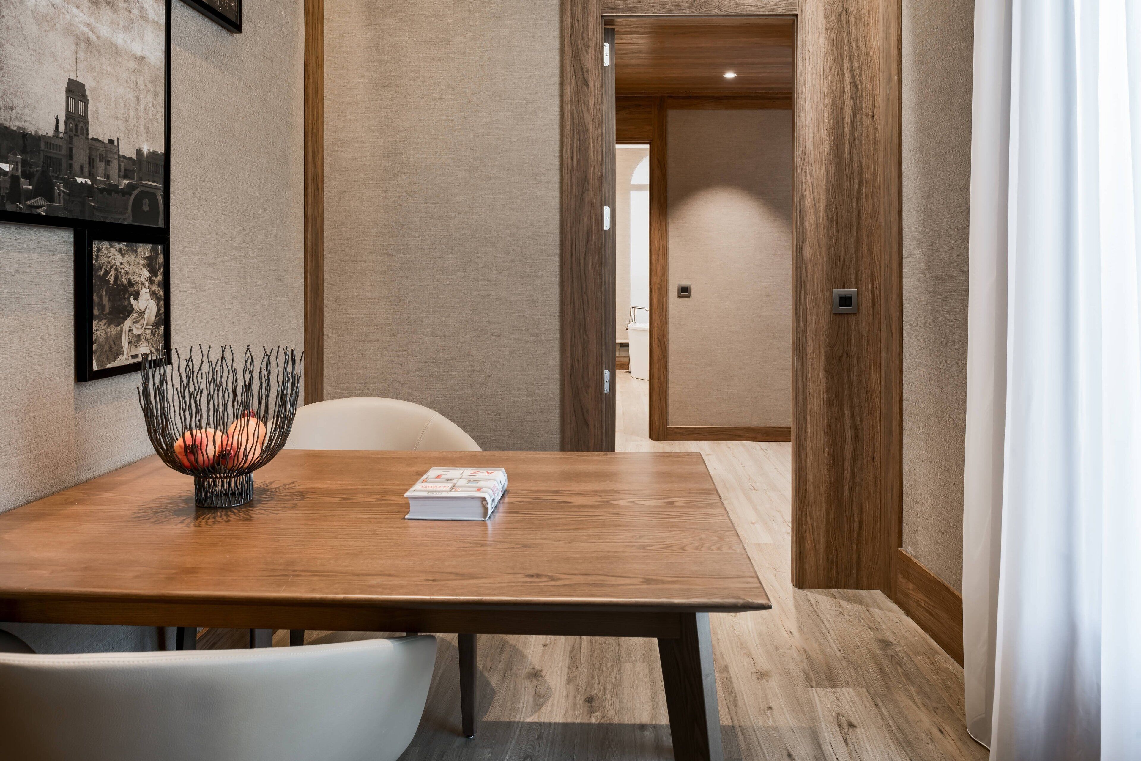 Un comedor con una mesa de madera, sillas blancas y un cuenco decorativo. Hay cuadros en la pared y una puerta que da a otra habitación.