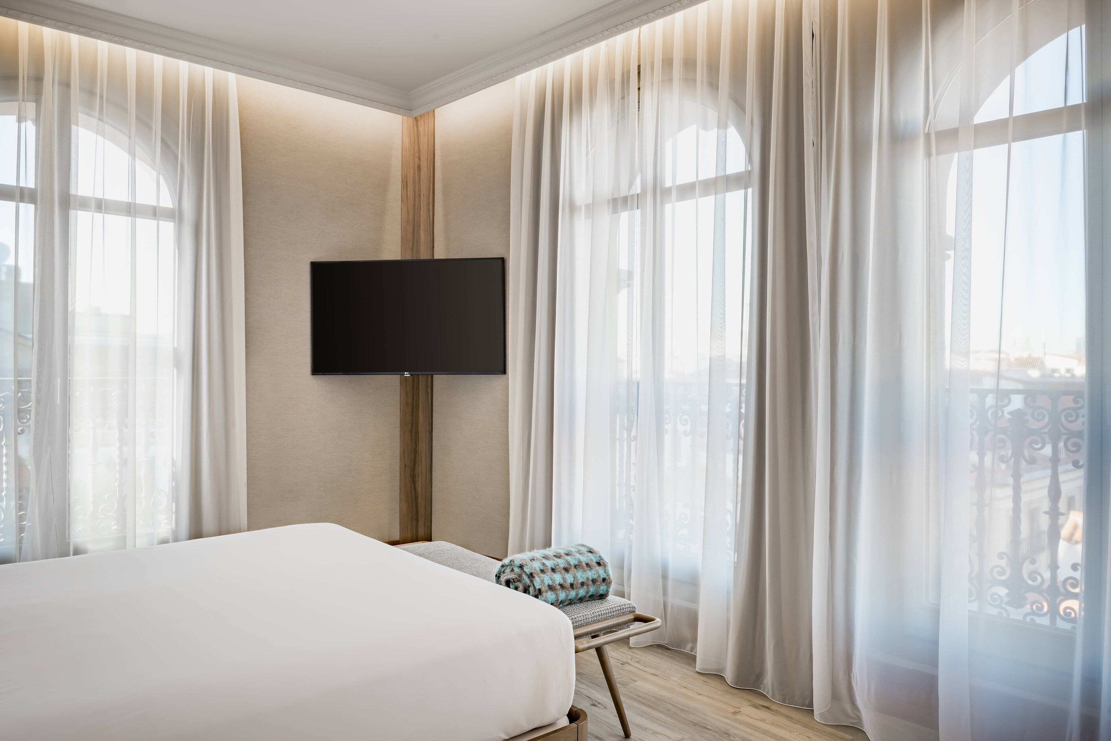 A hotel room with a large bed, a flat-screen TV mounted on the wall, and sheer curtains on the windows.
