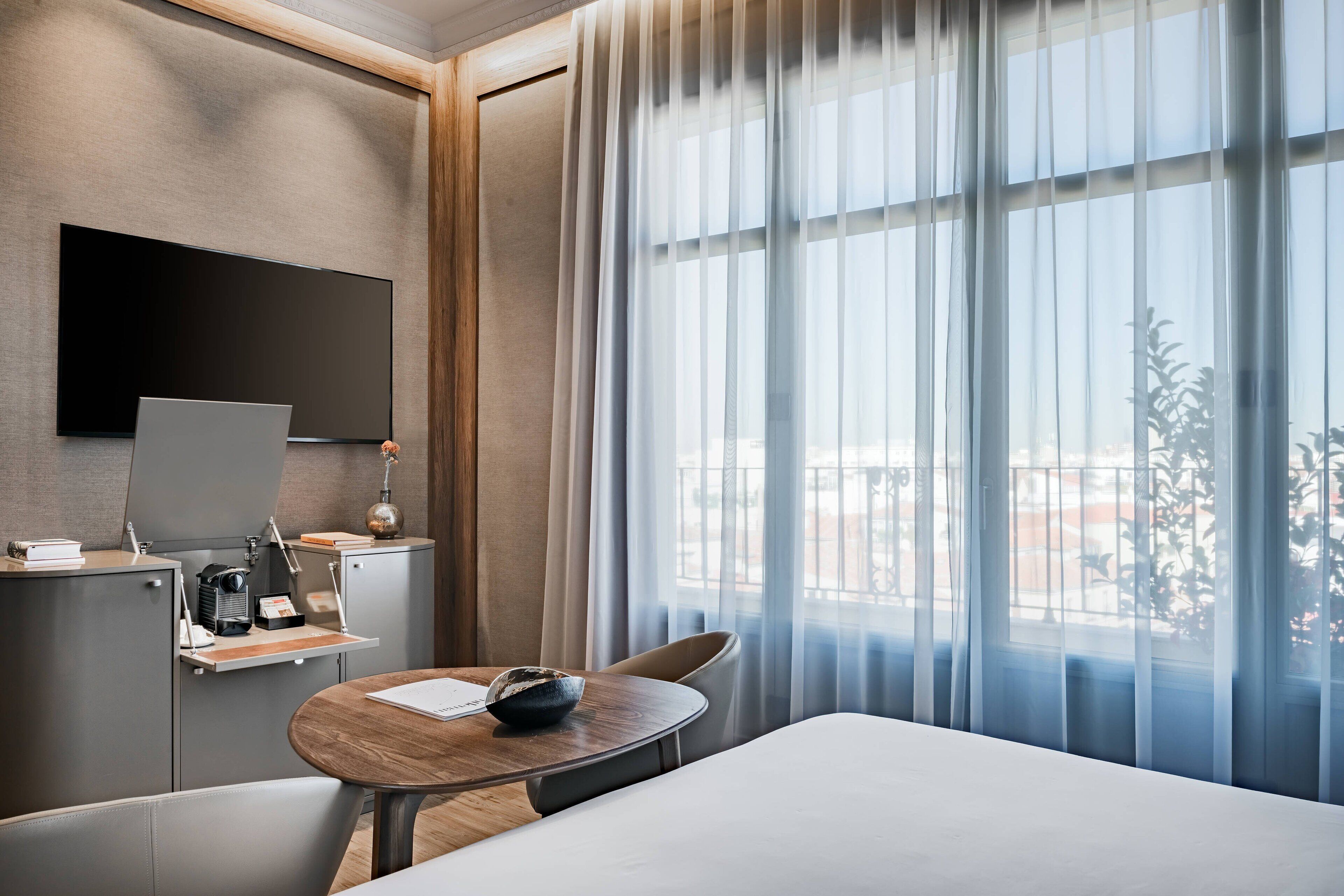 A modern hotel room with a bed, a desk with a flat-screen TV, a coffee maker, and a window with sheer curtains.
