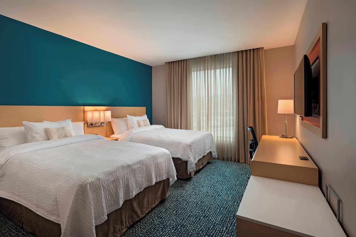 A hotel room with two beds, a desk, a TV, and a large window with curtains.