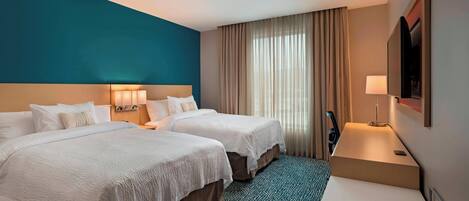 A hotel room with two beds, a desk, a TV, and a large window with curtains.