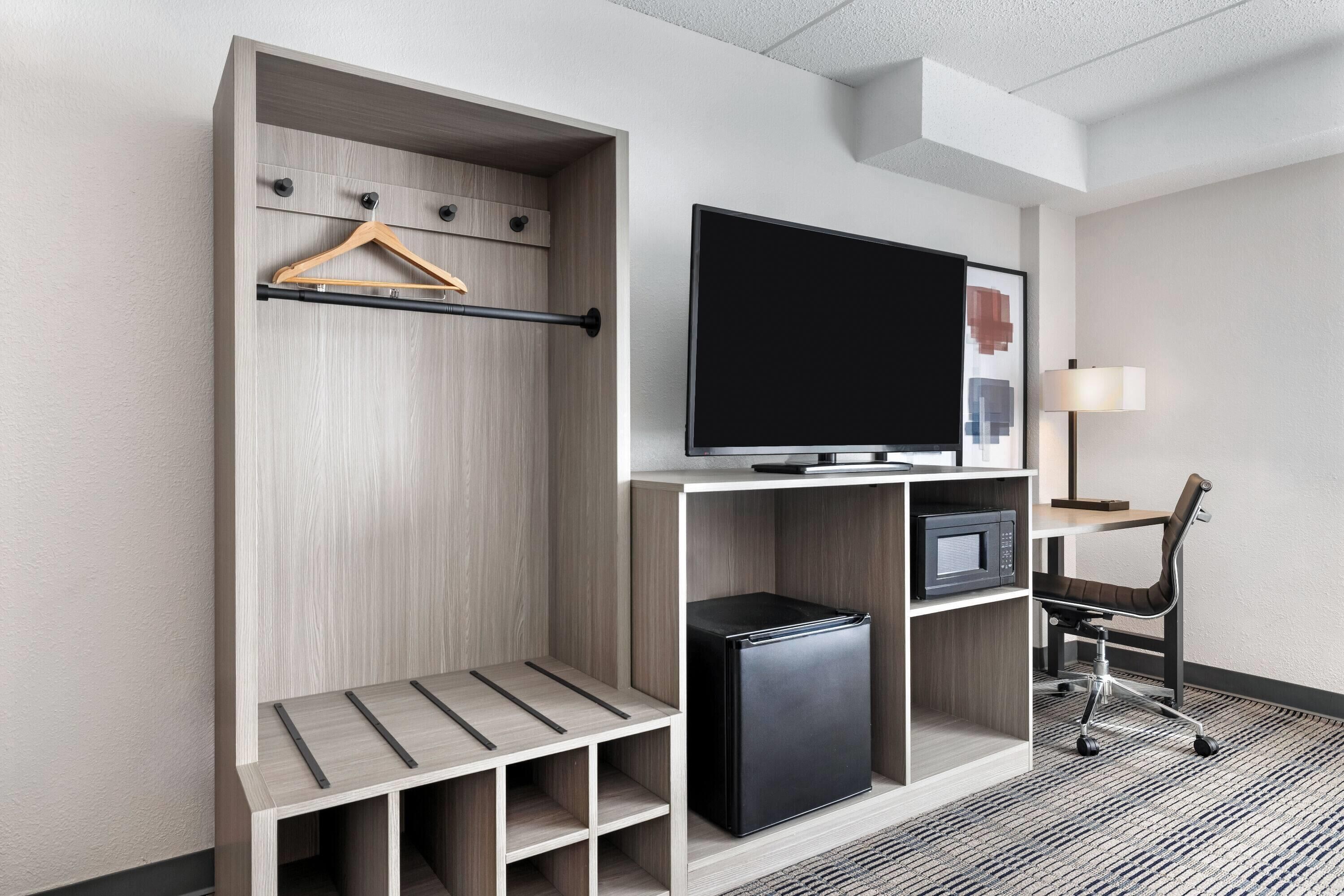 A modern hotel room with a built-in wardrobe, a flat-screen TV, a desk with a chair, and a small refrigerator.