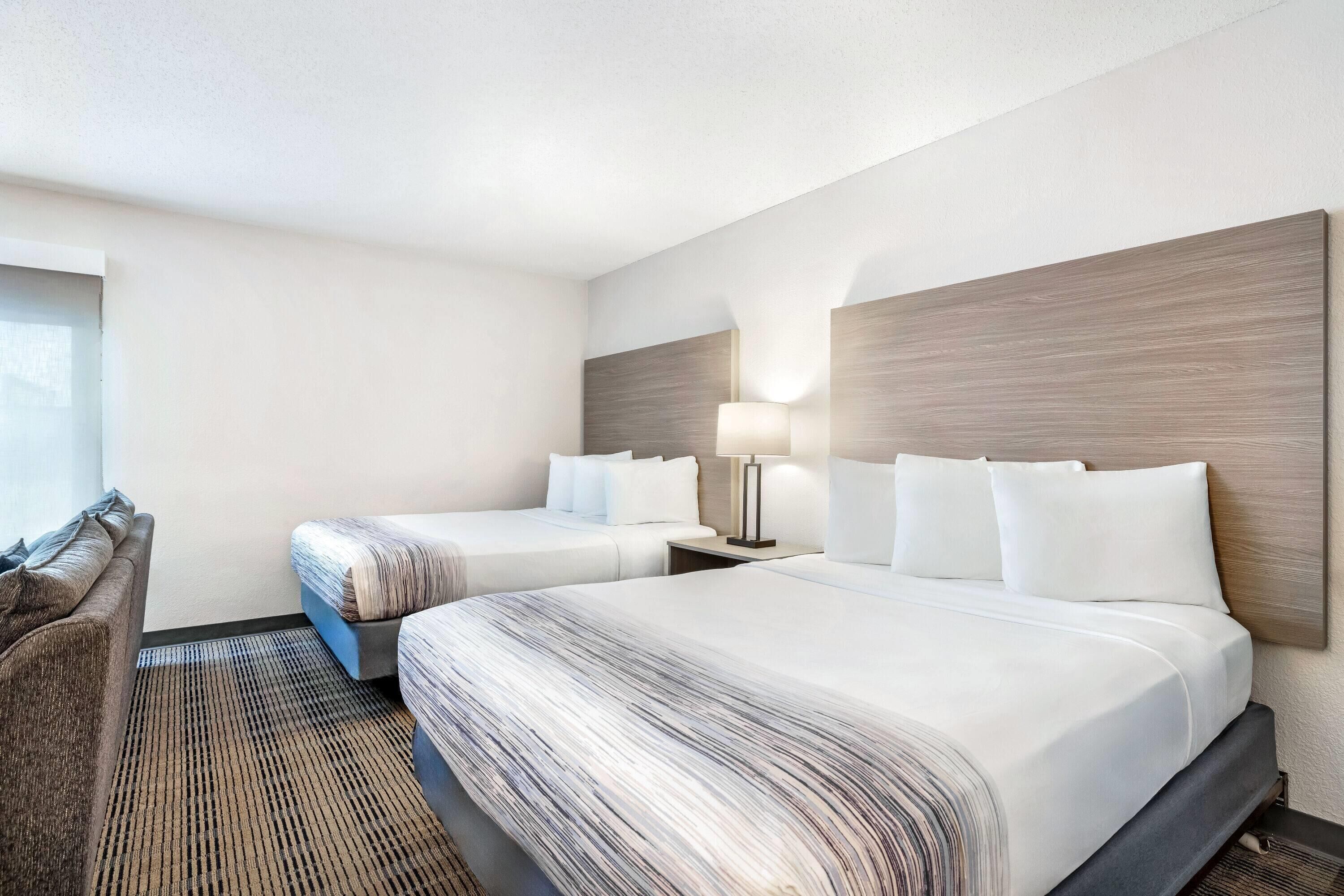 Quarto de hotel com duas camas, uma mesinha de cabeceira com abajur, uma cadeira e uma janela grande.