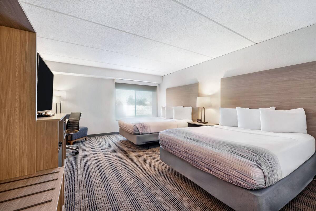 A hotel room with a large bed, a smaller bed, a desk with a chair, a window, and a lamp.