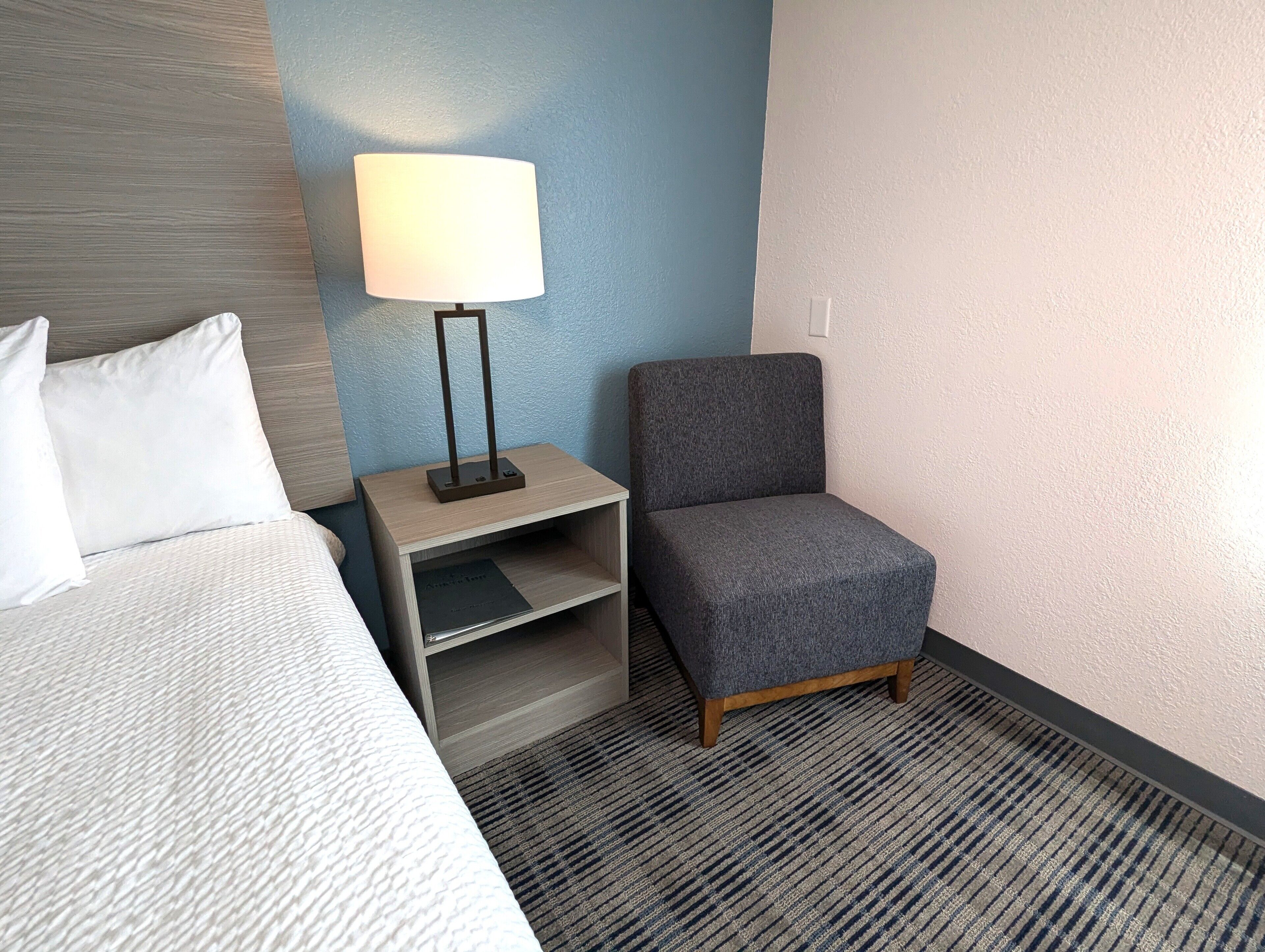 Une chambre d’hôtel avec un lit, une table de chevet avec une lampe, un fauteuil gris et un tapis à motifs.