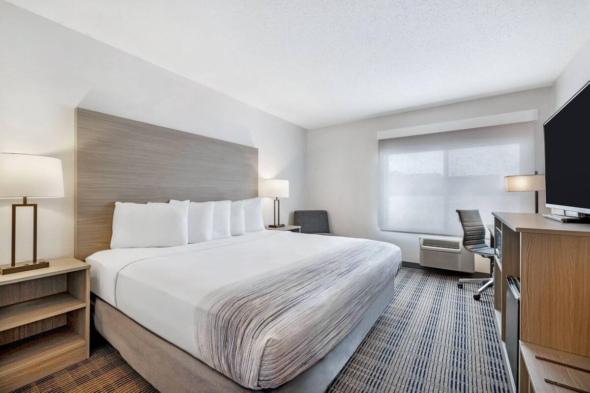 A hotel room with a large bed, two bedside tables with lamps, a desk with a chair, a television, and a window.