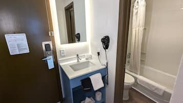 Une salle de bain équipée d’un lavabo, d’un miroir et de toilettes. Un plan d’évacuation en cas d’incendie est affiché sur la porte, ainsi qu’un sèche-cheveux et un porte-serviettes.