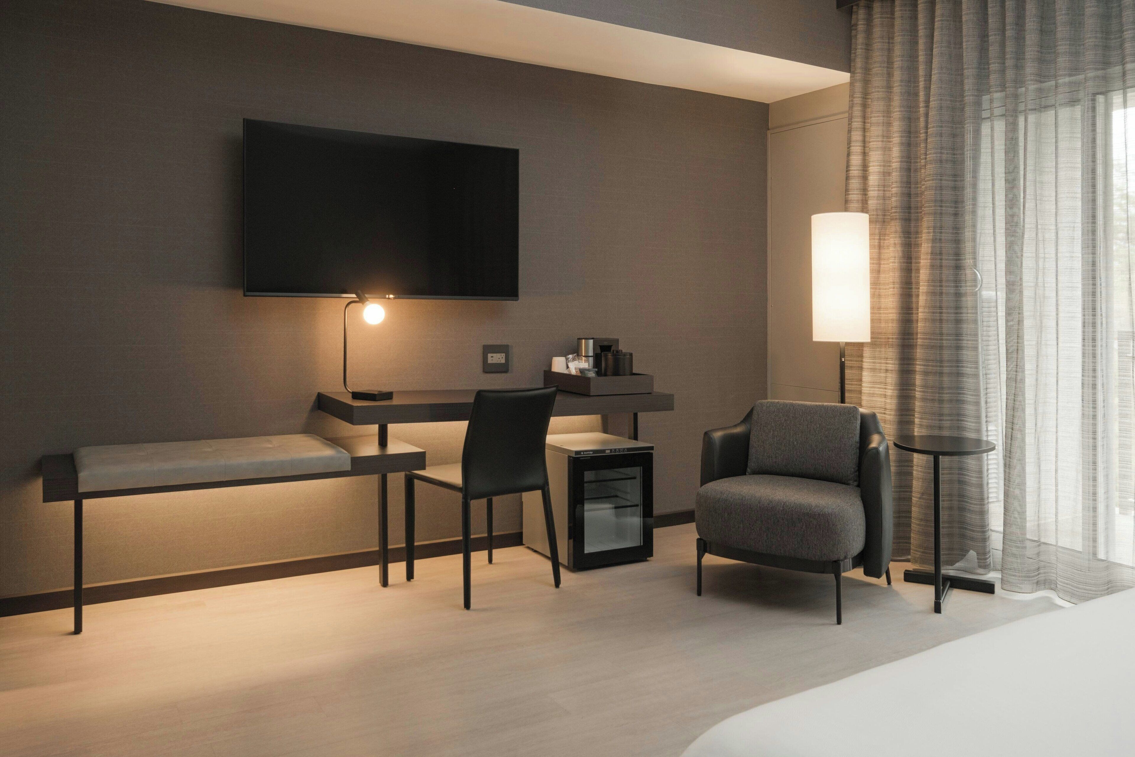 Une chambre d’hôtel moderne équipée d’un téléviseur à écran plat fixé au mur, d’un bureau avec une chaise, d’un petit réfrigérateur, d’un canapé et d’une table de chevet avec une lampe.